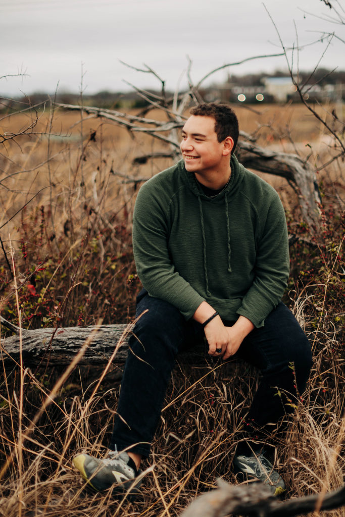 A senior from Harrison Arkansas sits on a tree log with dry grass around him and smiles as he looks at the horizon
